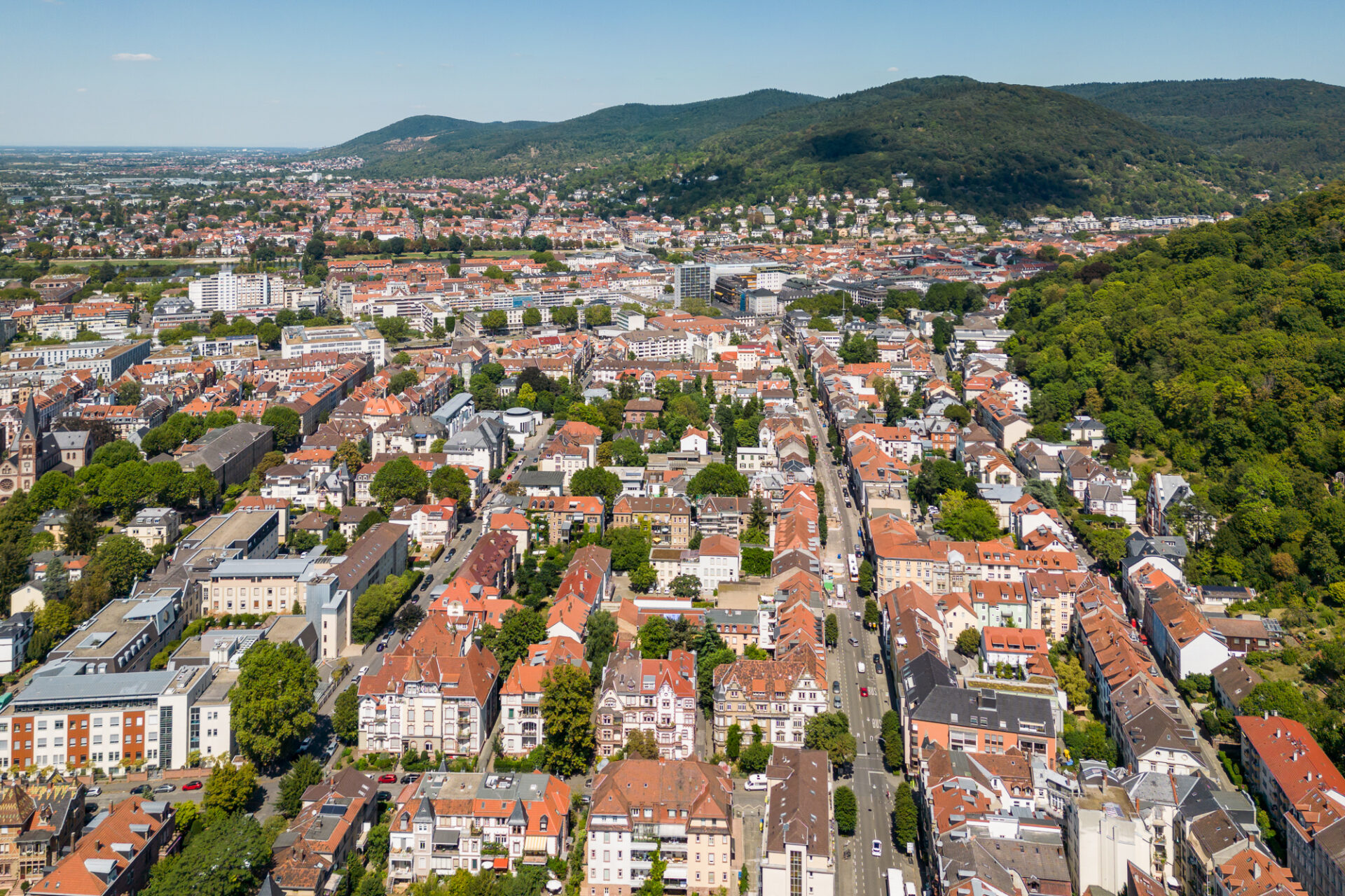 Luftaufnahme von Heidelberg, einer europäischen Stadt mit dichter Bebauung und grünen Hügeln im Hintergrund an einem sonnigen Tag.