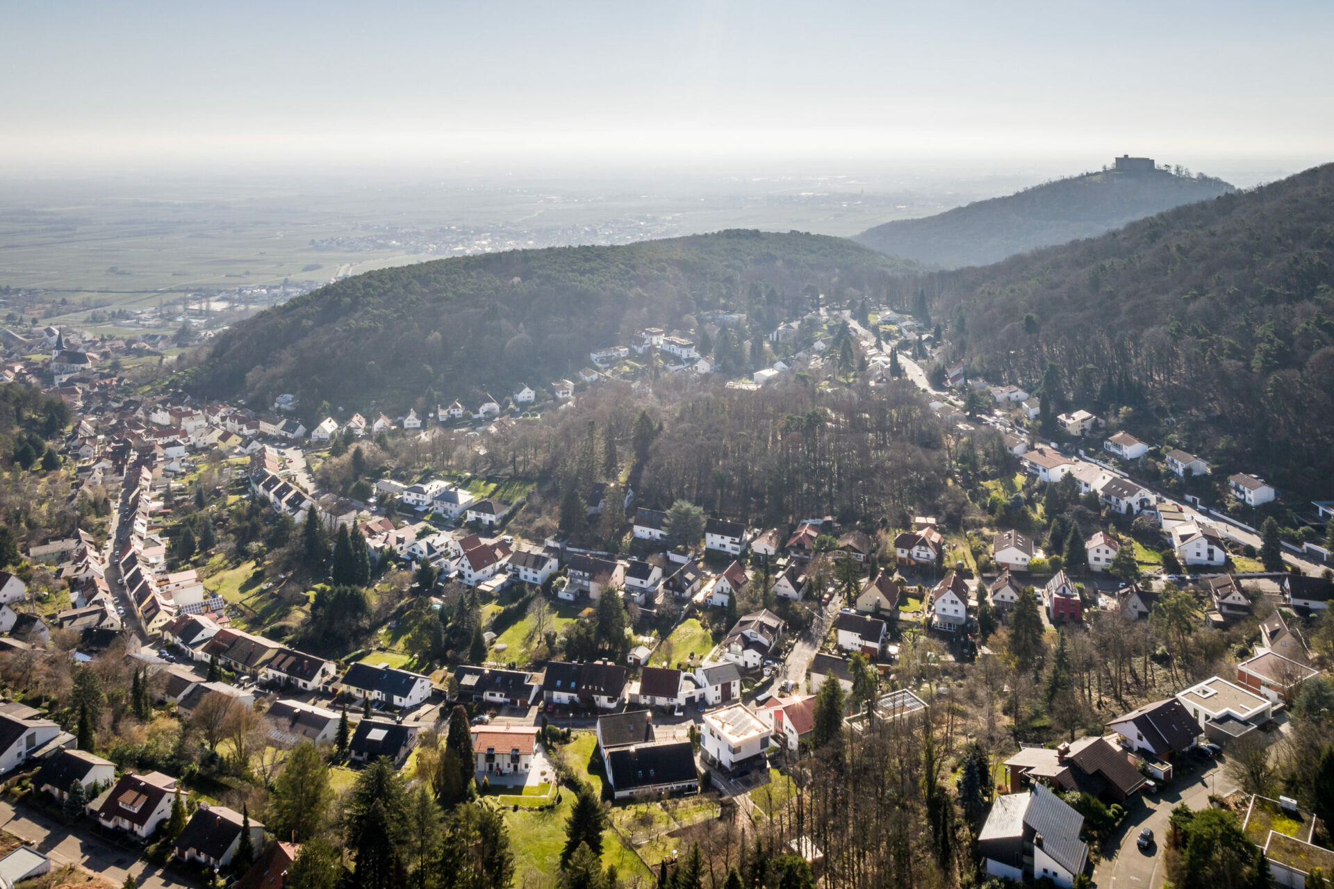 Luftaufnahme einer kleinen Stadt in einem Tal mit dem Hambacher Schloss auf einem nahe gelegenen Hügel, umgeben von üppigem Grün und klarem Himmel.