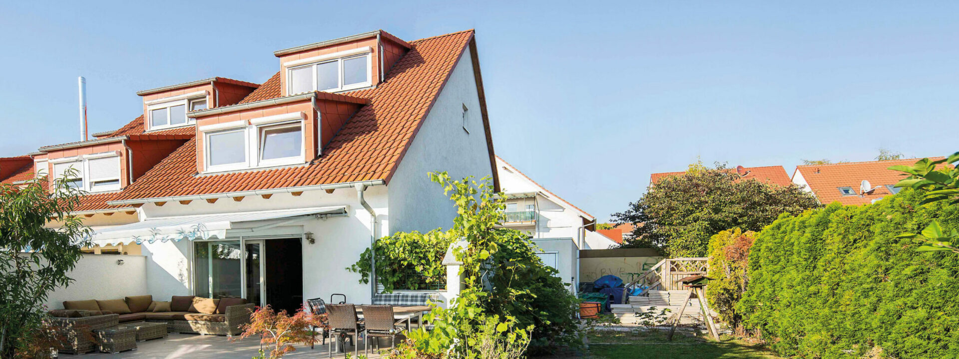 Ein modernes zweistöckiges Haus in Seckenheim mit weißer Fassade, rotem Dach und üppigem Garten unter einem klaren blauen Himmel.