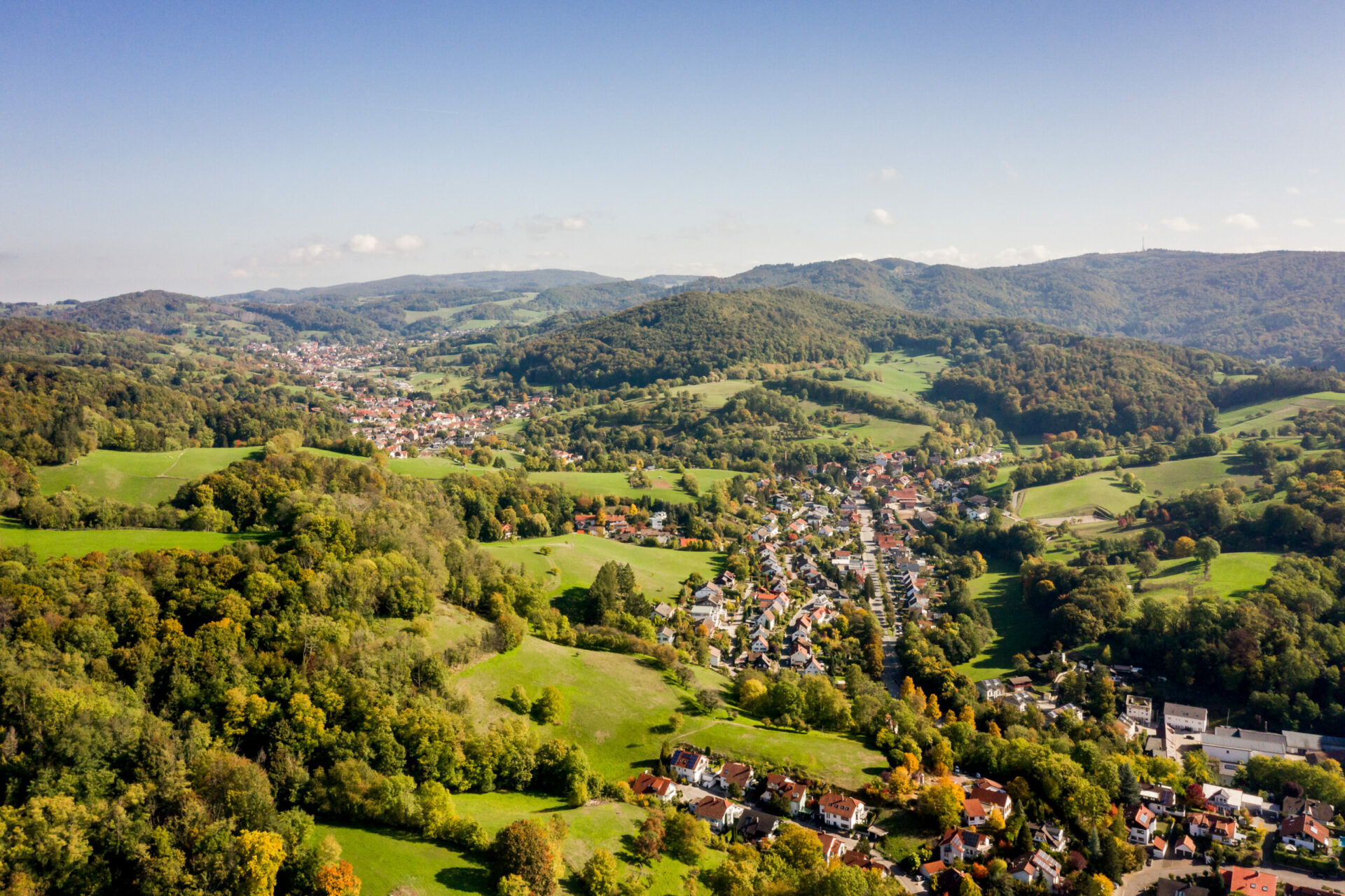 Luftaufnahme einer üppigen, grünen Landschaft mit der Kleinstadt Bensheim mit gruppierten Häusern, umgeben von sanften Hügeln und Wäldern unter einem klaren Himmel.