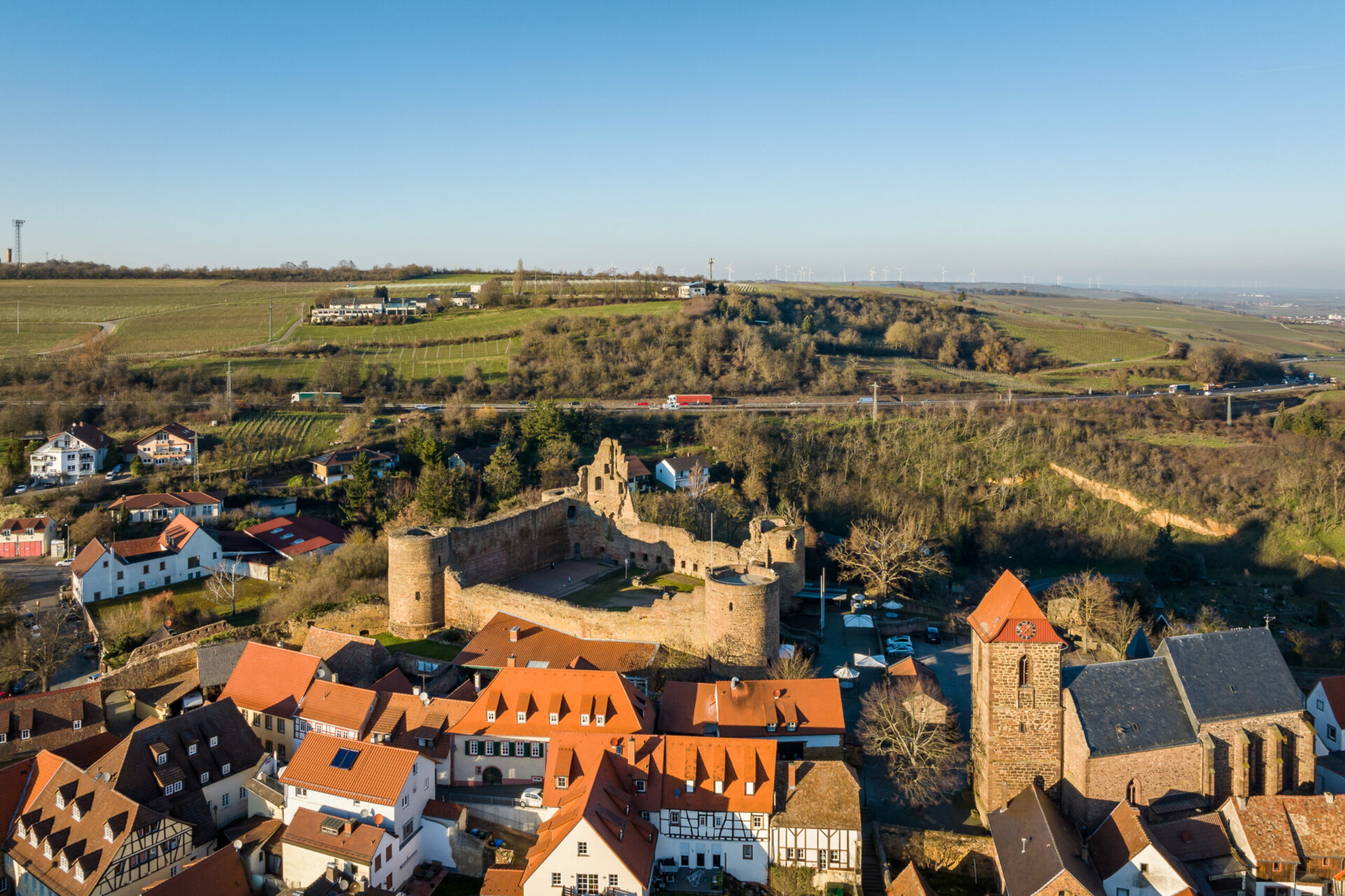 Luftaufnahme des deutschen Dorfes Neuleiningen mit mittelalterlichen Burgruinen, umgeben von modernen Häusern und üppigem Grün, unter einem klaren blauen Himmel.