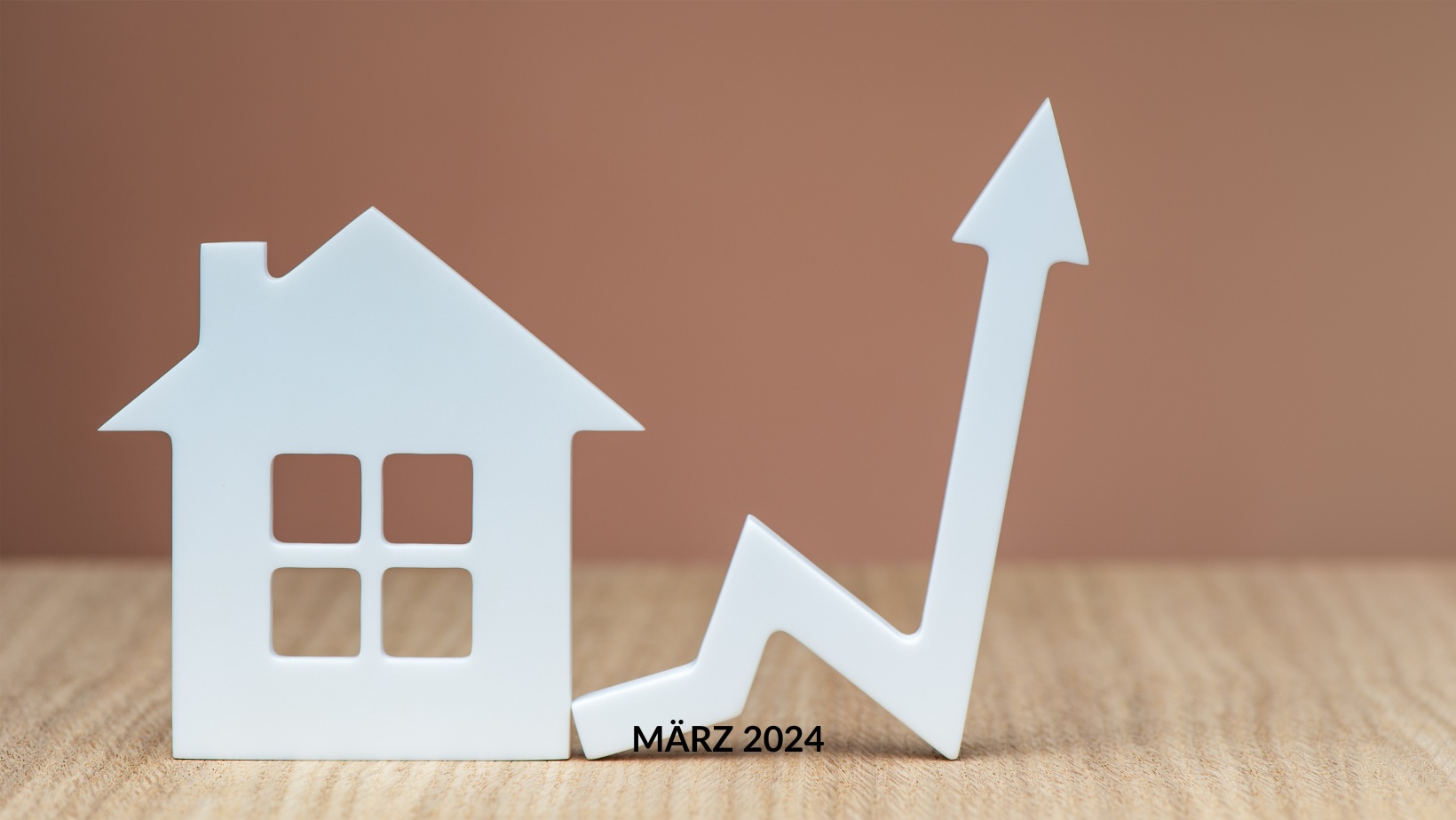 Weißer Ausschnitt eines Hauses neben einem nach oben gerichteten Pfeil mit der Aufschrift „März 2024“ auf einem Holztisch, symbolisiert Metropolen-Immobilienpreise-Trends oder Prognosen für März