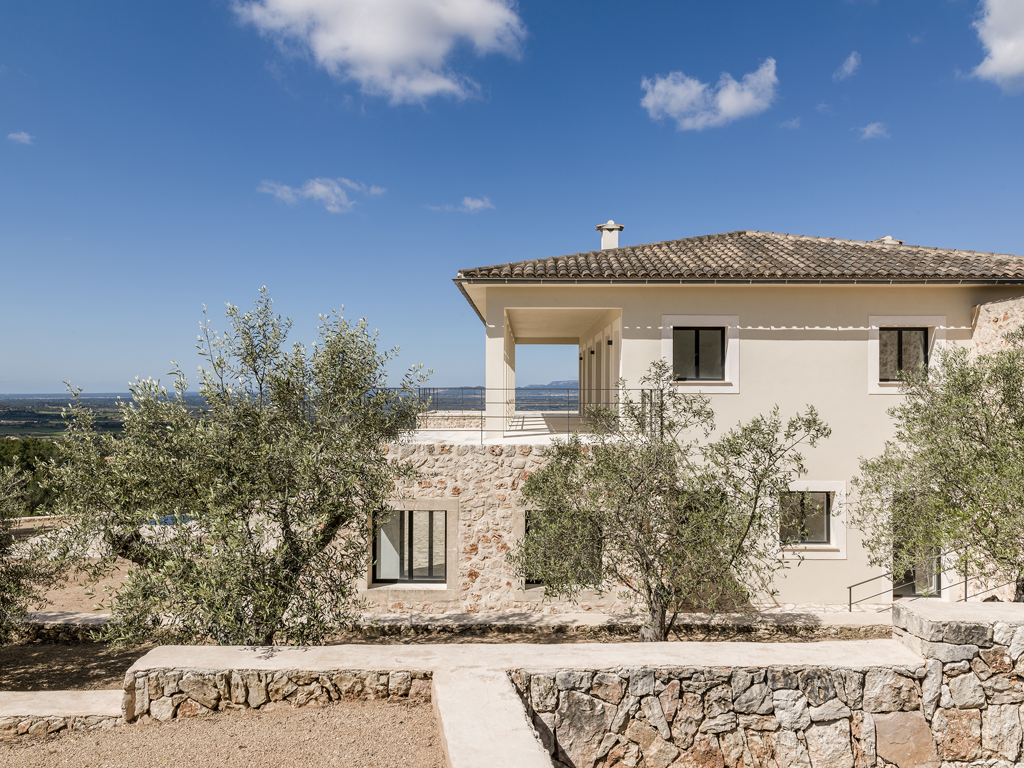 Zweistöckiges Haus mit Stein- und Gipsfassade, umgeben von Olivenbäumen unter einem klaren blauen Himmel, mit internationalen Angeboten.